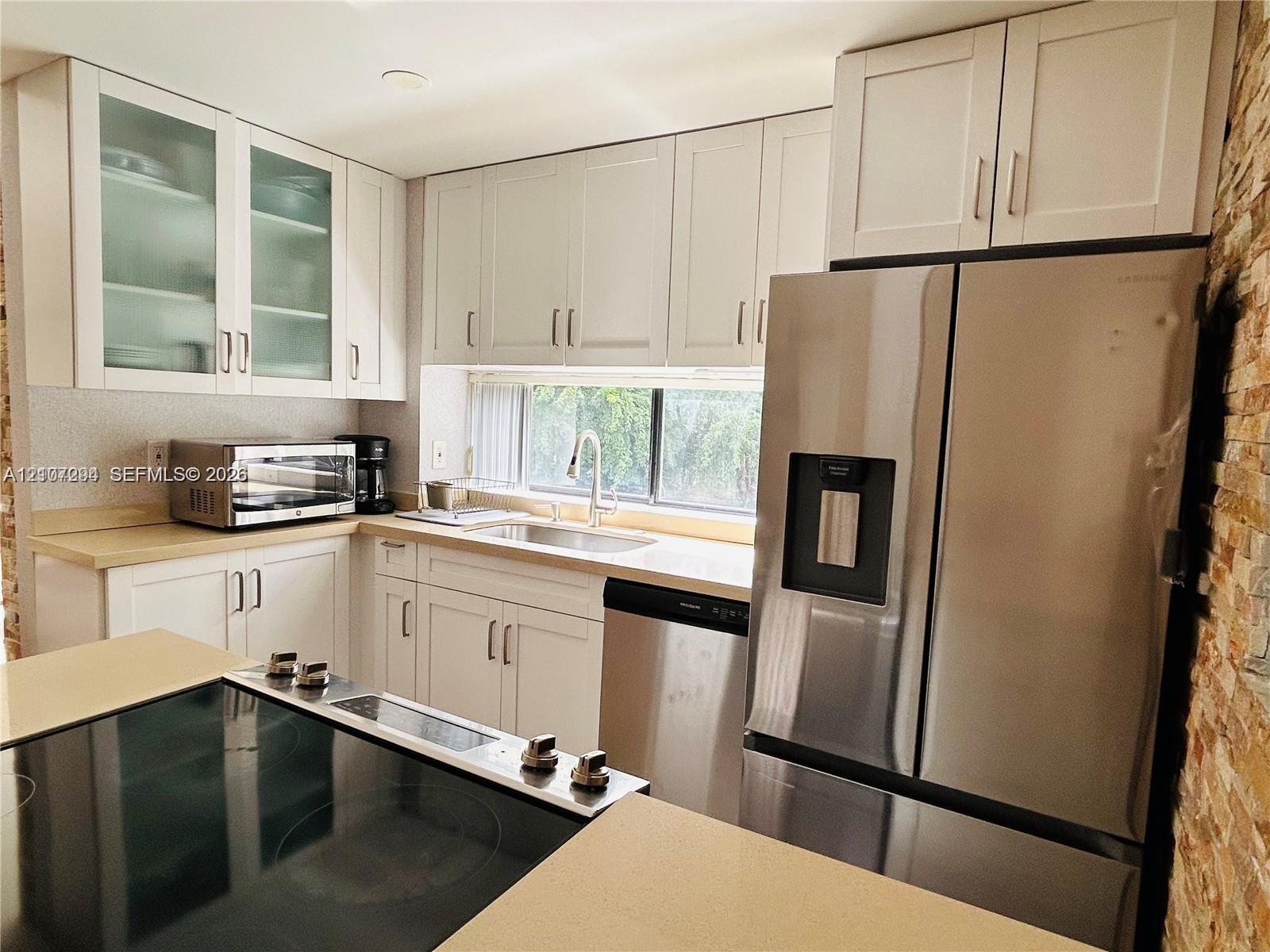 Kitchen featuring white Shaker-style cabinetry with glass-front uppers, stainless steel appliances, a stone-textured accent wall, and a solid-surface countertop with an integrated cooktop