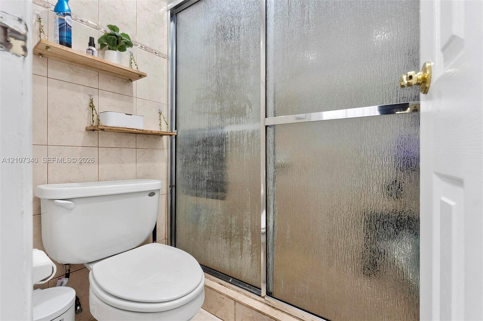 Bathroom featuring floor-to-ceiling neutral ceramic tile, a glass shower enclosure with metal framing, and wall-mounted shelving