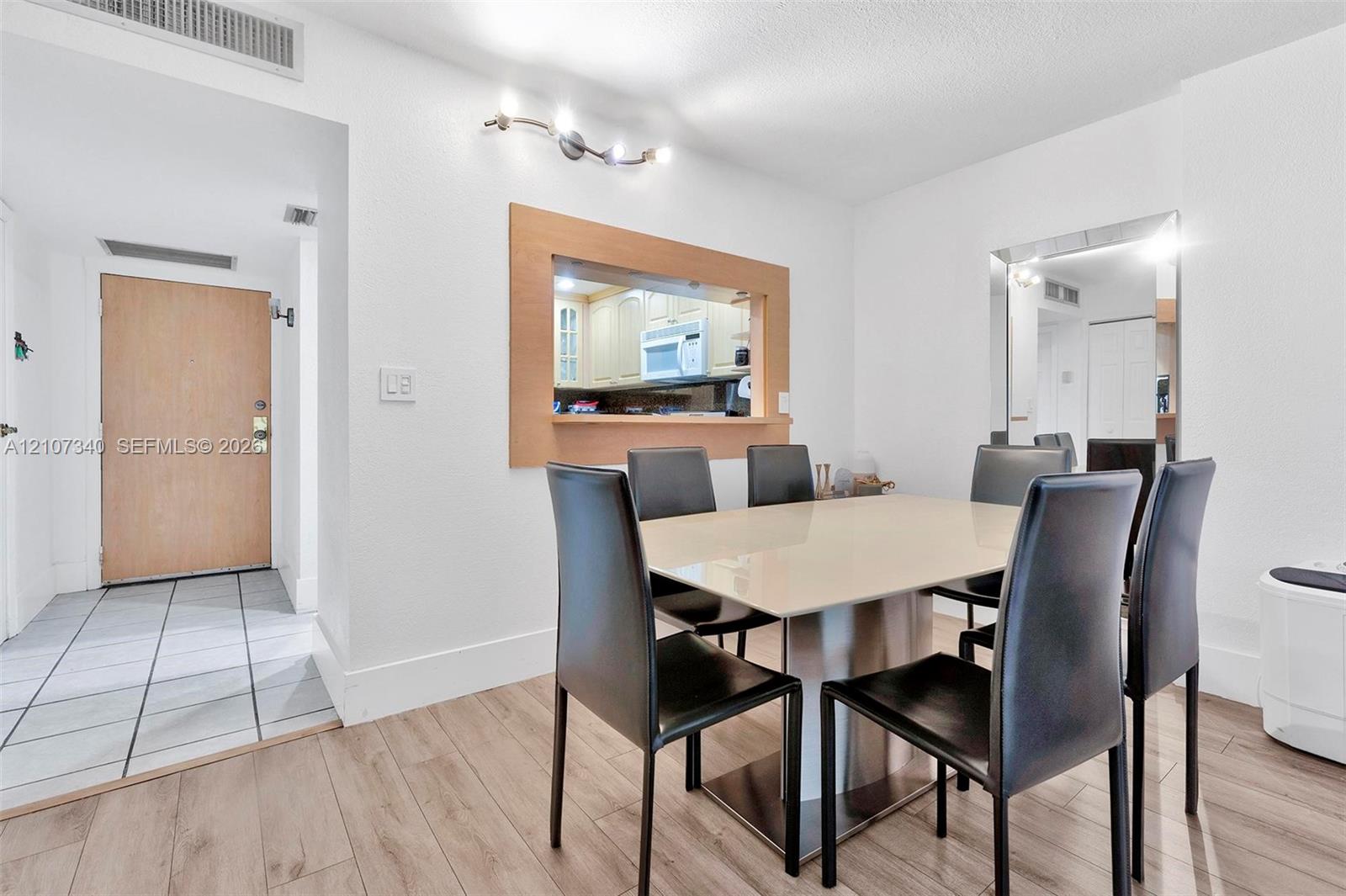 Dining area featuring wood-finish flooring, a wall-mounted multi-bulb light fixture, a framed pass-through opening to the kitchen, and a floor-to-ceiling mirror
