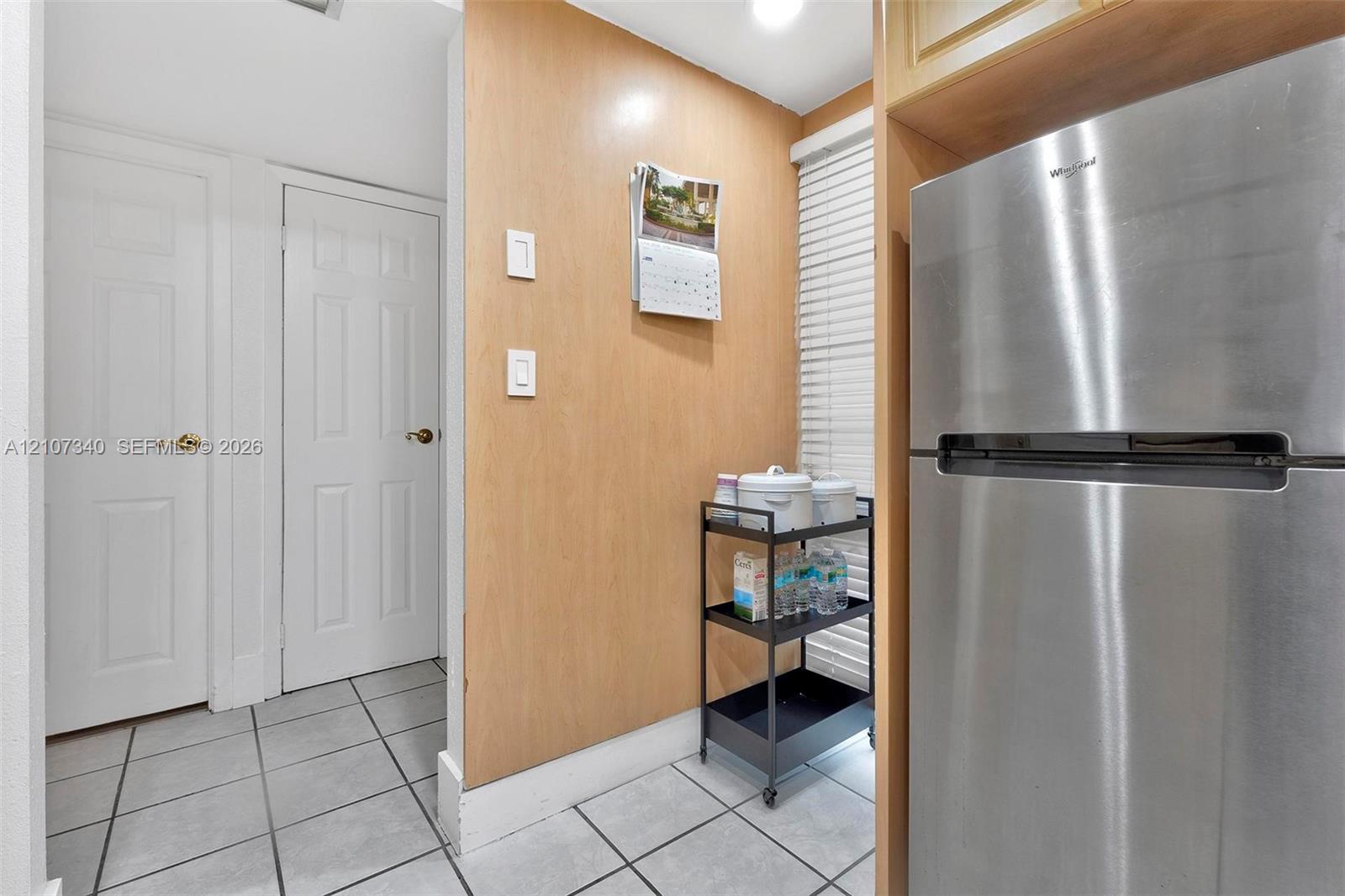 Stainless steel refrigerator, light-toned wood-finish wall paneling, white six-panel doors with metallic hardware, and ceramic tile flooring