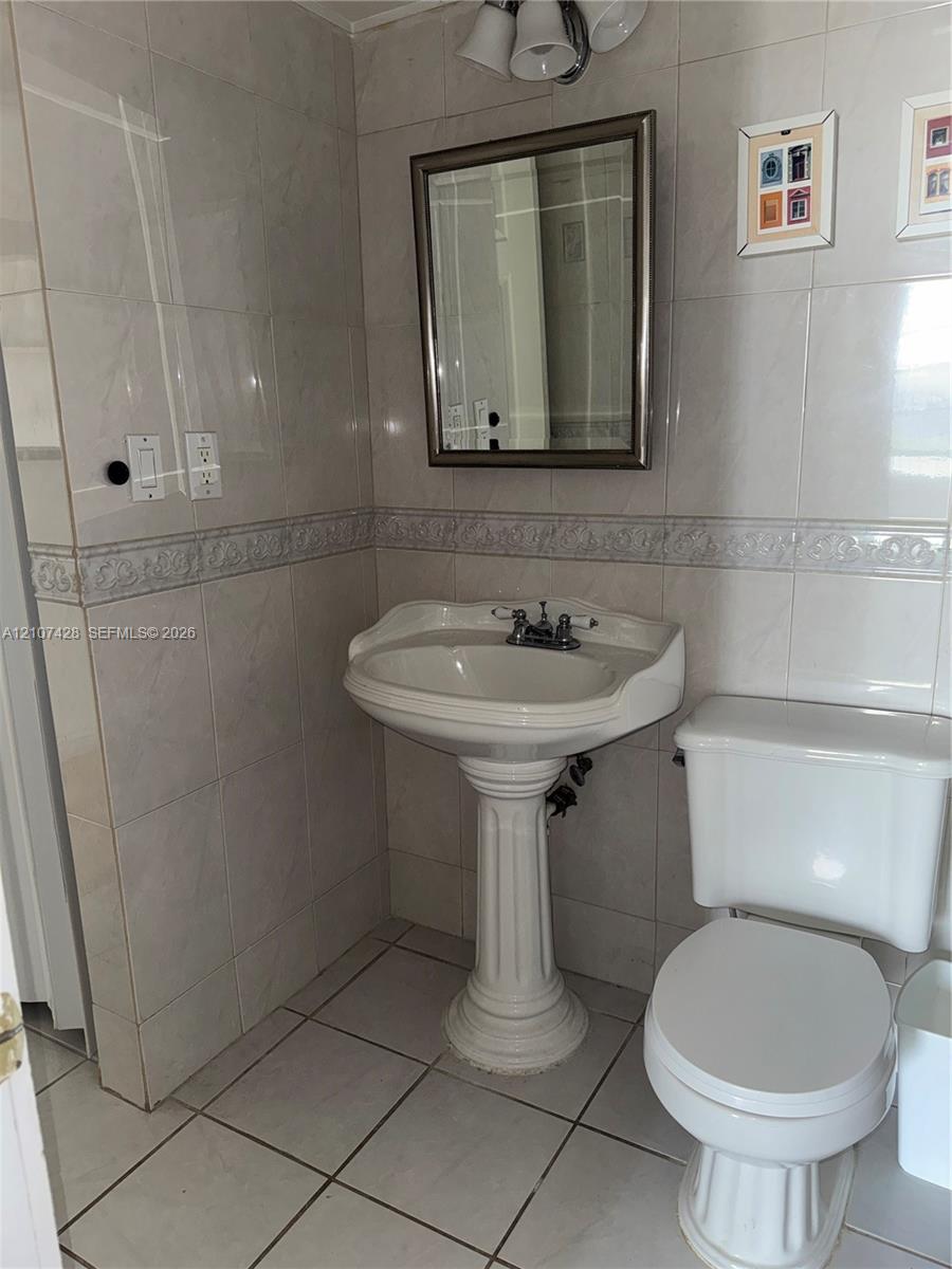 Full-wall tile surround with decorative accent border, white pedestal sink, polished chrome plumbing fixtures, rectangular wall-mounted mirror, and tile flooring