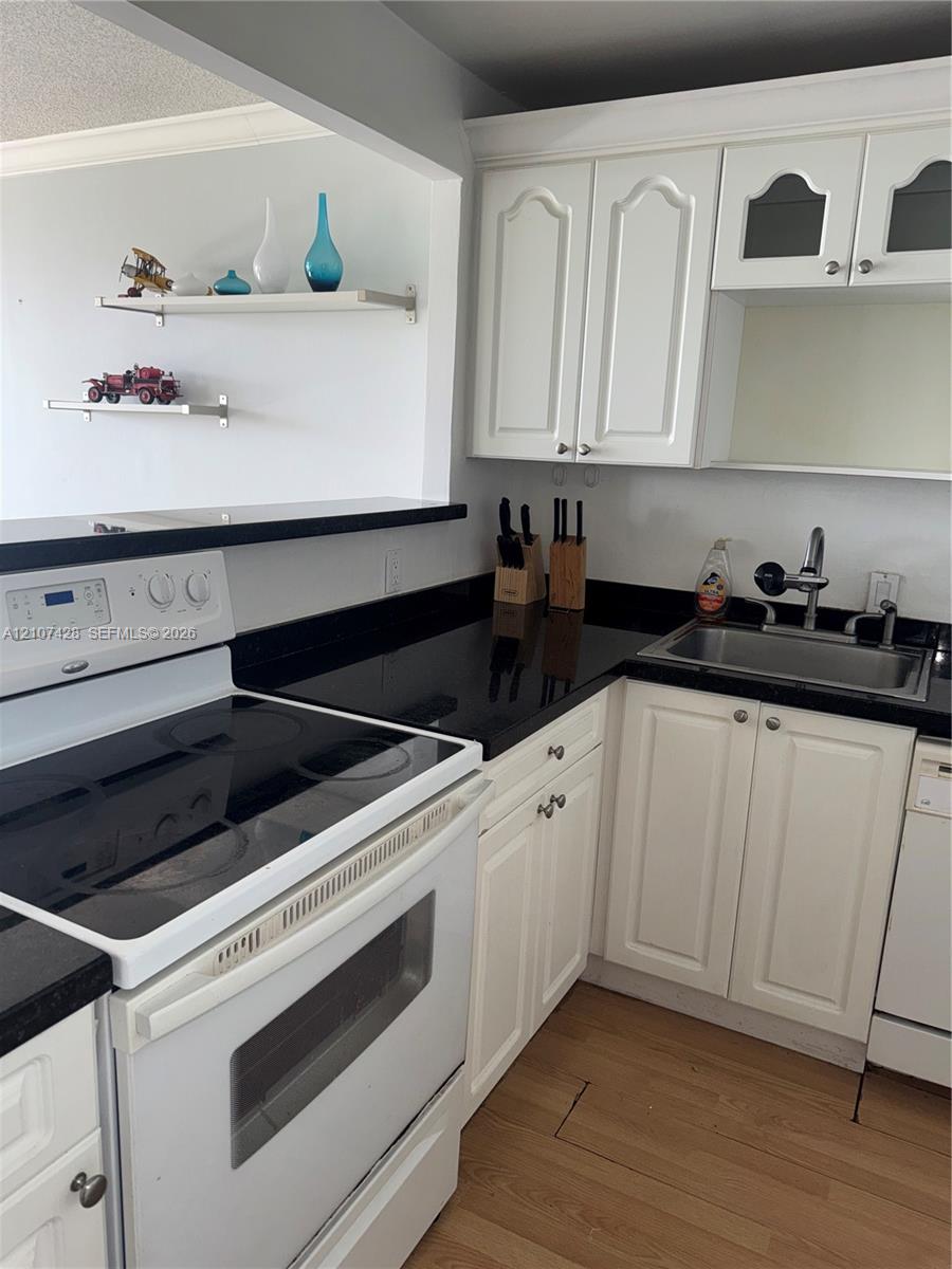 Kitchen featuring white cabinetry with glass-panel accents, dark countertops, wood-finish flooring, two wall-mounted floating shelves, and an electric range