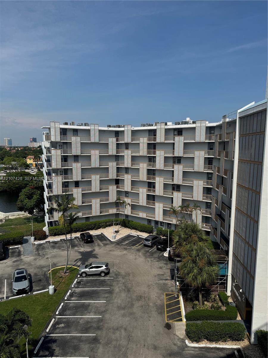 Mid-rise residential building featuring a structural geometric facade, stacked private balconies, dedicated surface parking, and mature palm landscaping