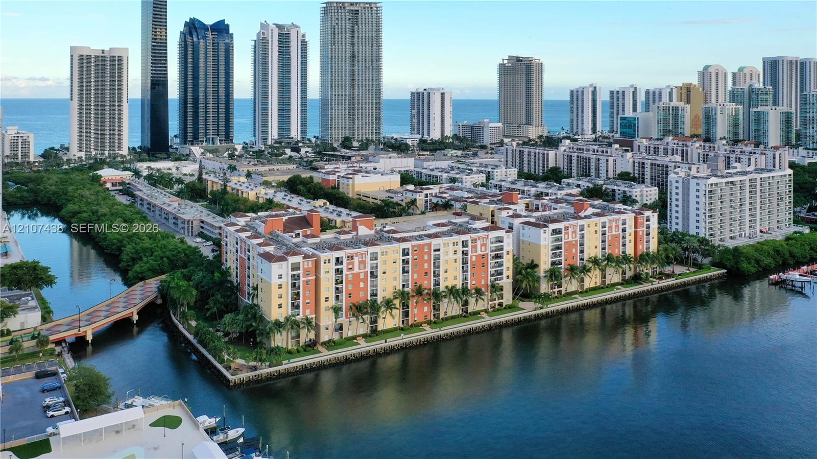 Aerial perspective of a multi-story residential complex situated along a waterway, featuring balcony access, palm tree landscaping, and proximity to the coastline