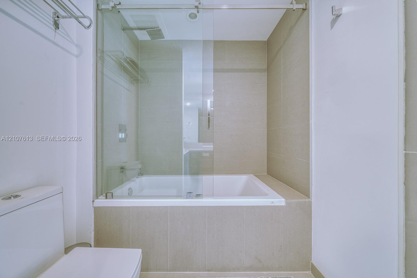 Soaking tub with a glass sliding enclosure, floor-to-ceiling neutral tile surround, and wall-mounted metal fixtures