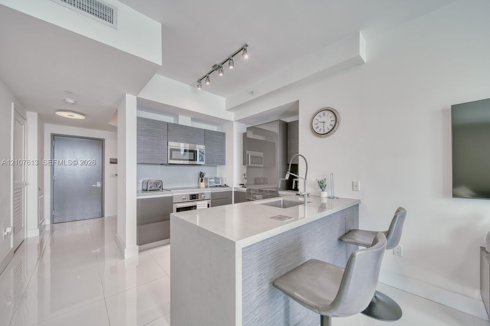 Kitchen featuring flat-panel grey cabinetry, quartz countertops, integrated stainless steel appliances, a breakfast bar with a high-arc faucet, and polished tile flooring