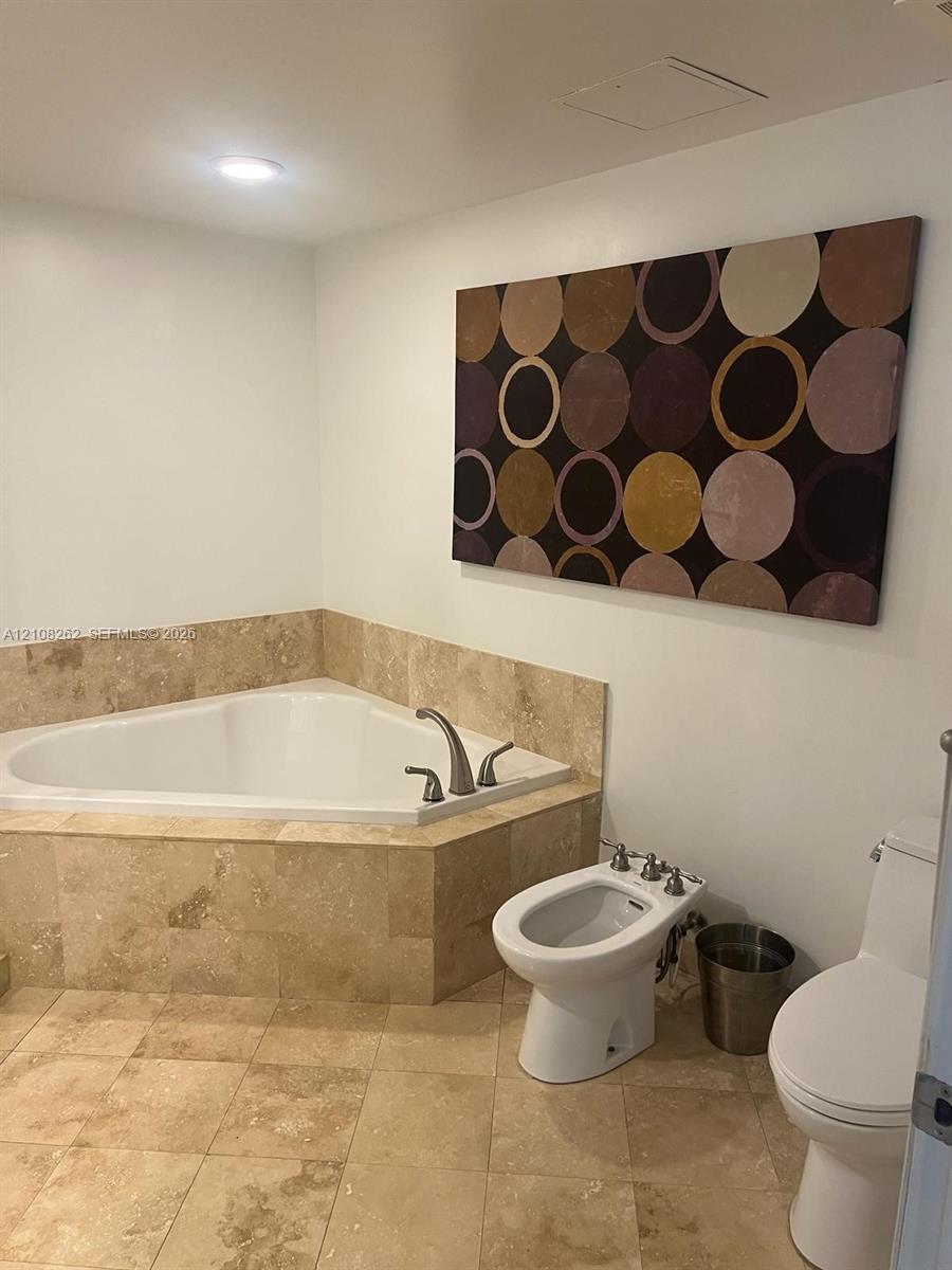 Corner soaking tub with natural stone surround, stone tile flooring, bidet, and two-piece toilet