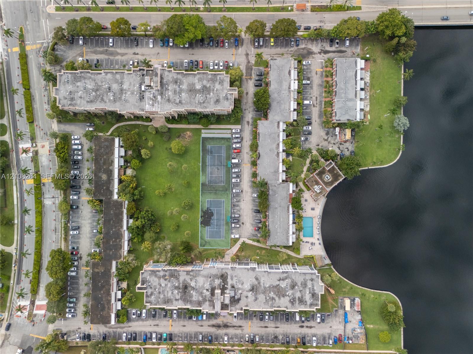 Aerial perspective of the residential complex featuring a swimming pool, two tennis courts, expansive green space, and waterfront frontage