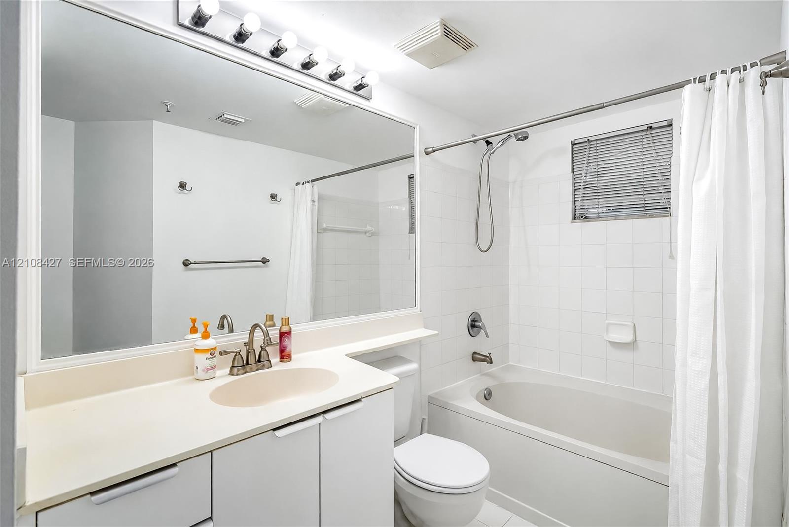 Bathroom featuring a full-width vanity mirror, a stone-surfaced countertop with an integrated basin, a combination tub and shower with tile surround, and a four-bulb vanity light fixture