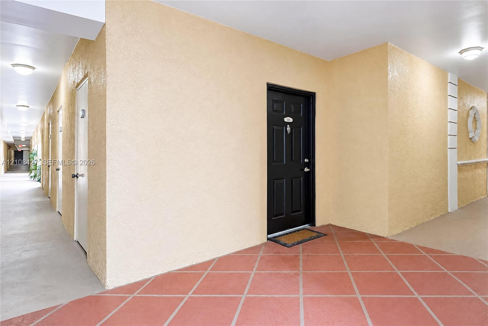 Entrance featuring a black six-panel door with metallic hardware, set against cream-colored textured walls with flush-mount ceiling lighting and terracotta-toned tile flooring