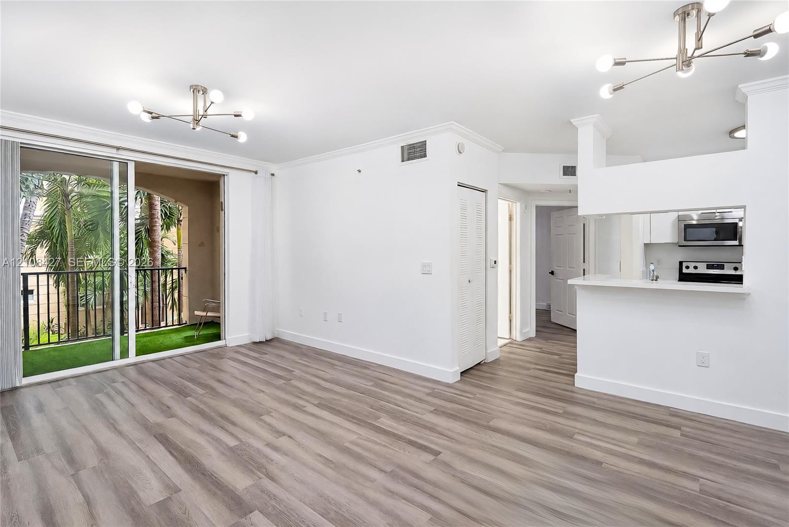Open living area featuring wood-finish flooring, crown molding, modern multi-arm light fixtures, a kitchen pass-through window with a breakfast bar, and sliding glass doors to a covered balcony