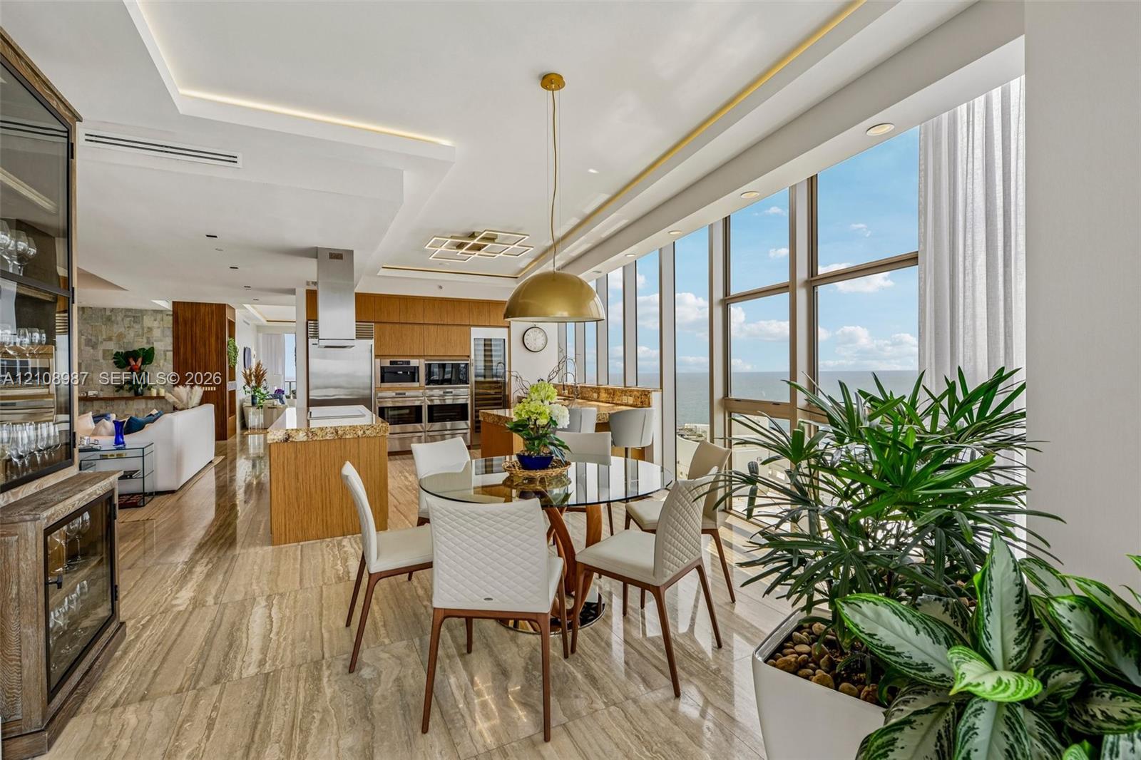 Open-concept space featuring floor-to-ceiling windows, polished stone flooring, custom wood-finish cabinetry, built-in stainless steel appliances, and a gold-tone pendant lighting fixture
