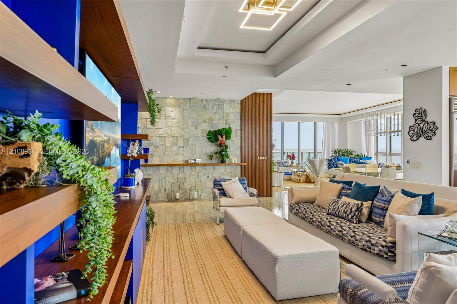 Custom built-in wood shelving with integrated bold blue accents, floor-to-ceiling stone-clad wall, recessed tray ceiling with geometric LED lighting, and large windows with natural illumination