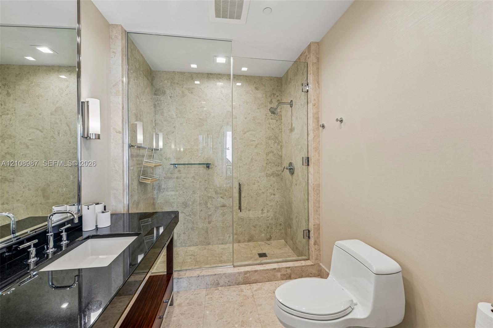 Bathroom featuring a wood-finish vanity with a dark polished countertop, undermount sink, walk-in shower with stone surround and frameless glass door, and recessed lighting