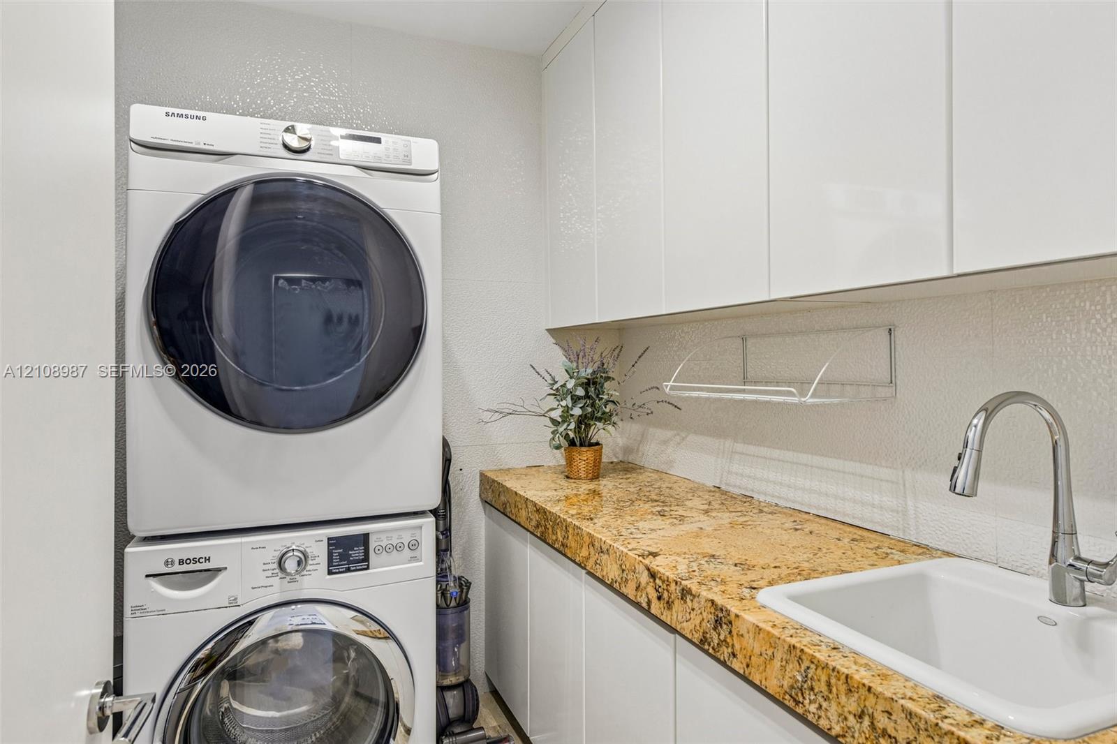 Laundry area featuring stacked appliances, stone-surface countertop, white gloss cabinetry, undermount sink, and chrome gooseneck faucet