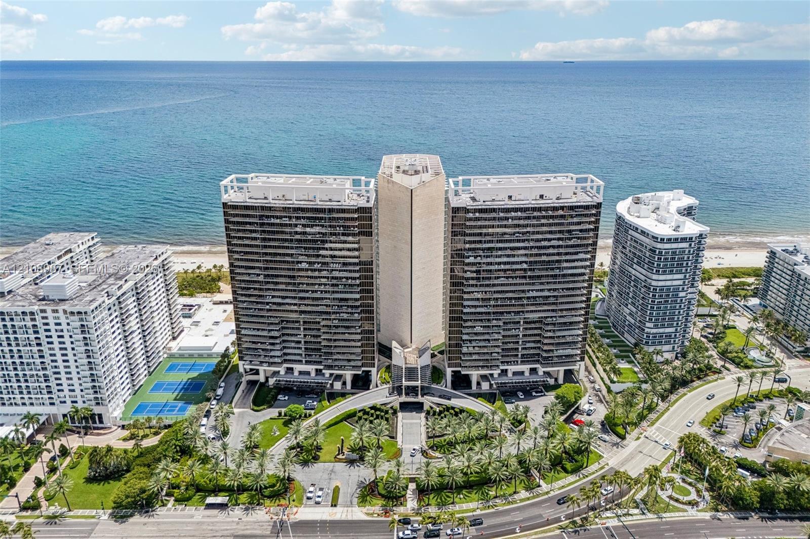 High-rise residential building featuring expansive ocean frontage, private balconies, on-site tennis courts, and landscaped grounds with mature palm trees