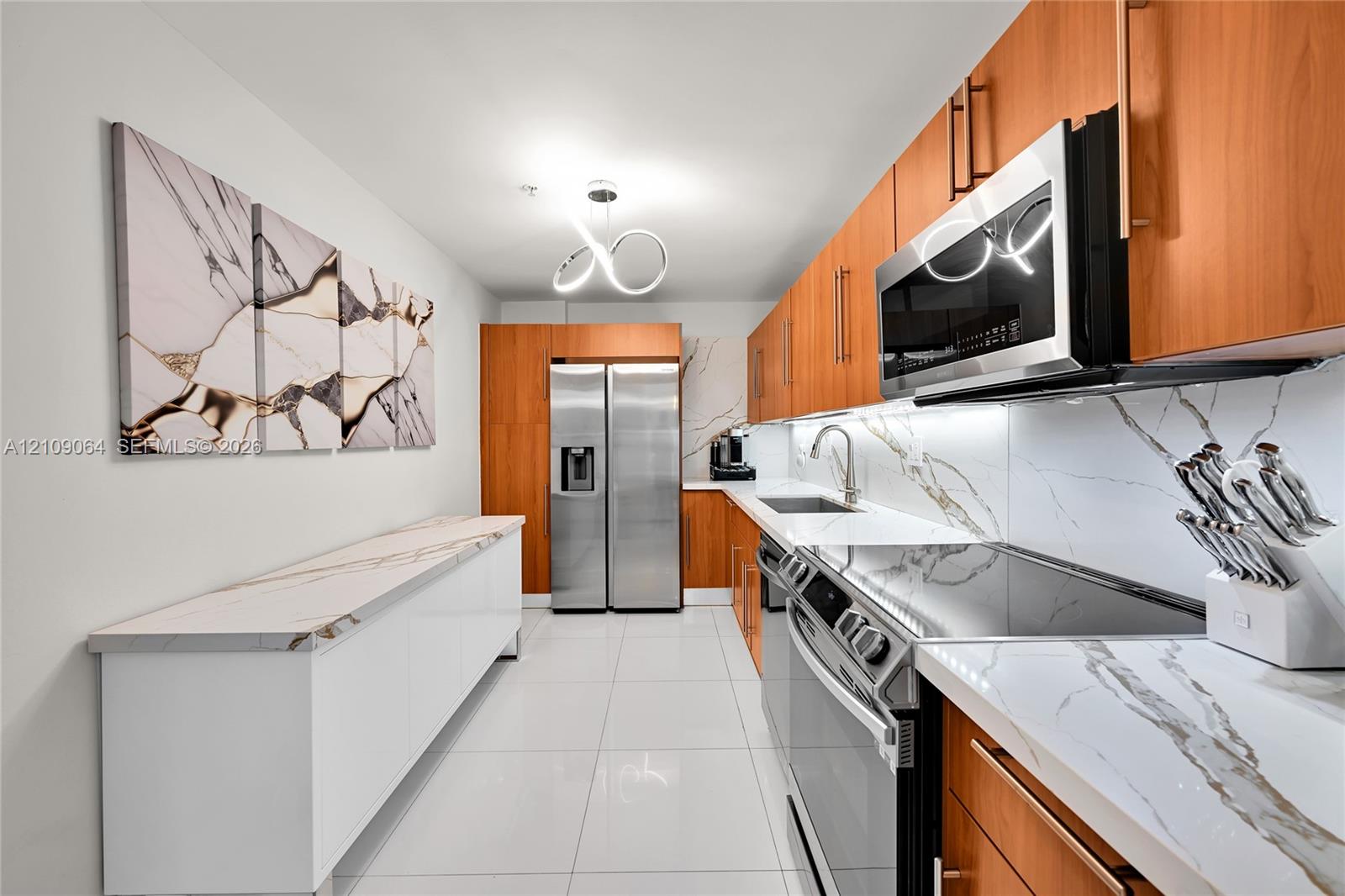 Kitchen featuring wood-finish cabinetry, stone-patterned backsplash, stainless steel appliances, under-cabinet lighting, and large-format tile flooring