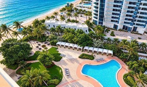 Aerial perspective of oceanfront property featuring a multi-level swimming pool, expansive paved terrace, linear cabanas, and manicured grounds