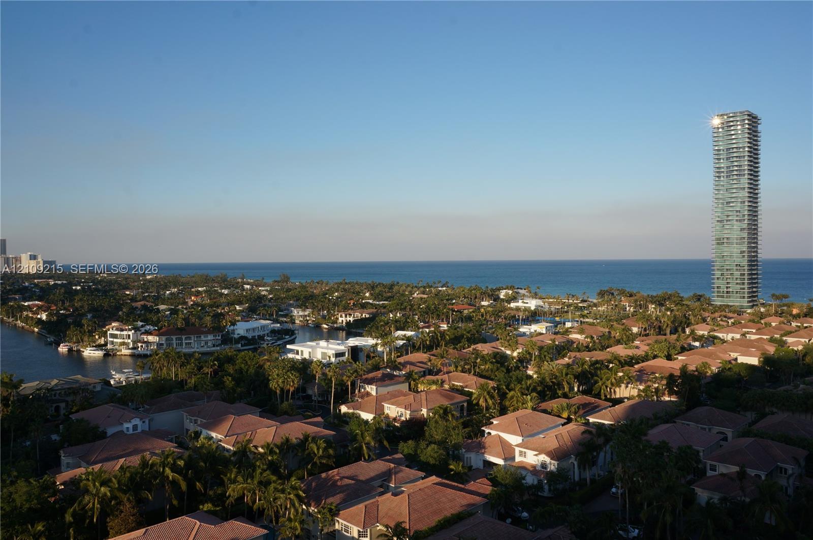 Aerial perspective featuring residential rooftops, mature vegetation, waterway access, and a coastal horizon