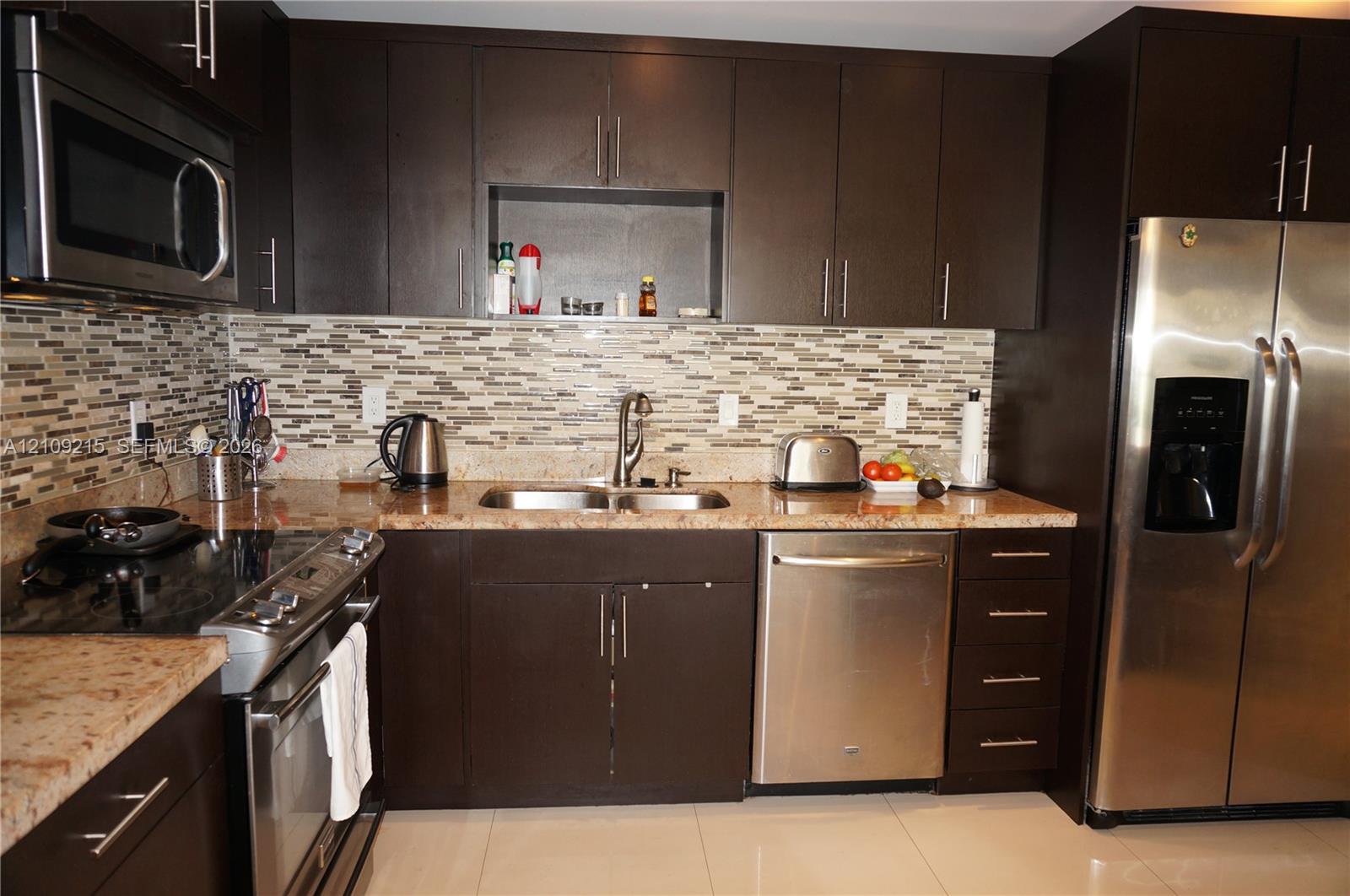 Dark espresso cabinetry with brushed metal pulls, stone countertops, horizontal multi-tonal backsplash, stainless steel appliances, and large-format tile flooring