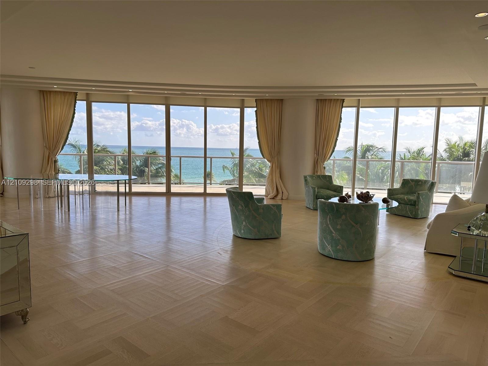 Expansive interior featuring parquet wood flooring, floor-to-ceiling curved glass wall panels, recessed lighting, and a metal-railed balcony with ocean scenery