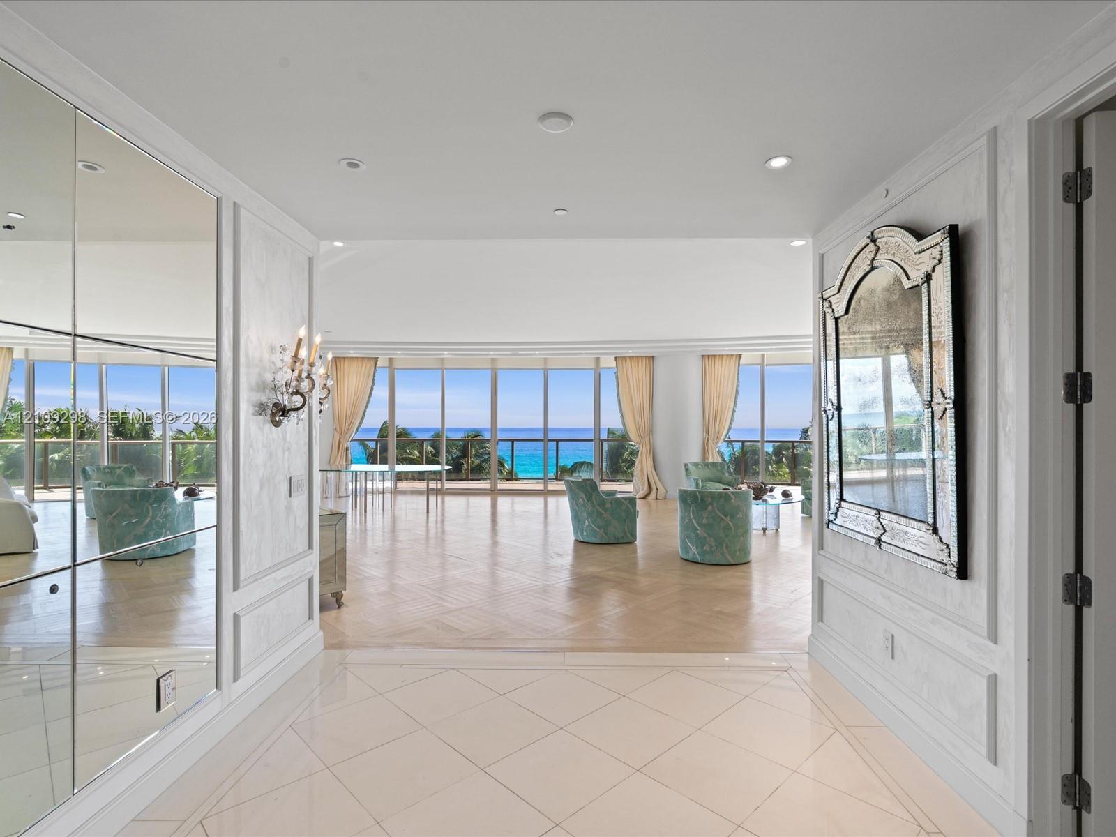 Entryway featuring large-format tile flooring, floor-to-ceiling mirrored wall panels, decorative wall molding, and recessed lighting, leading into a spacious room with herringbone wood-finish flooring and floor-to-ceiling glass panel windows