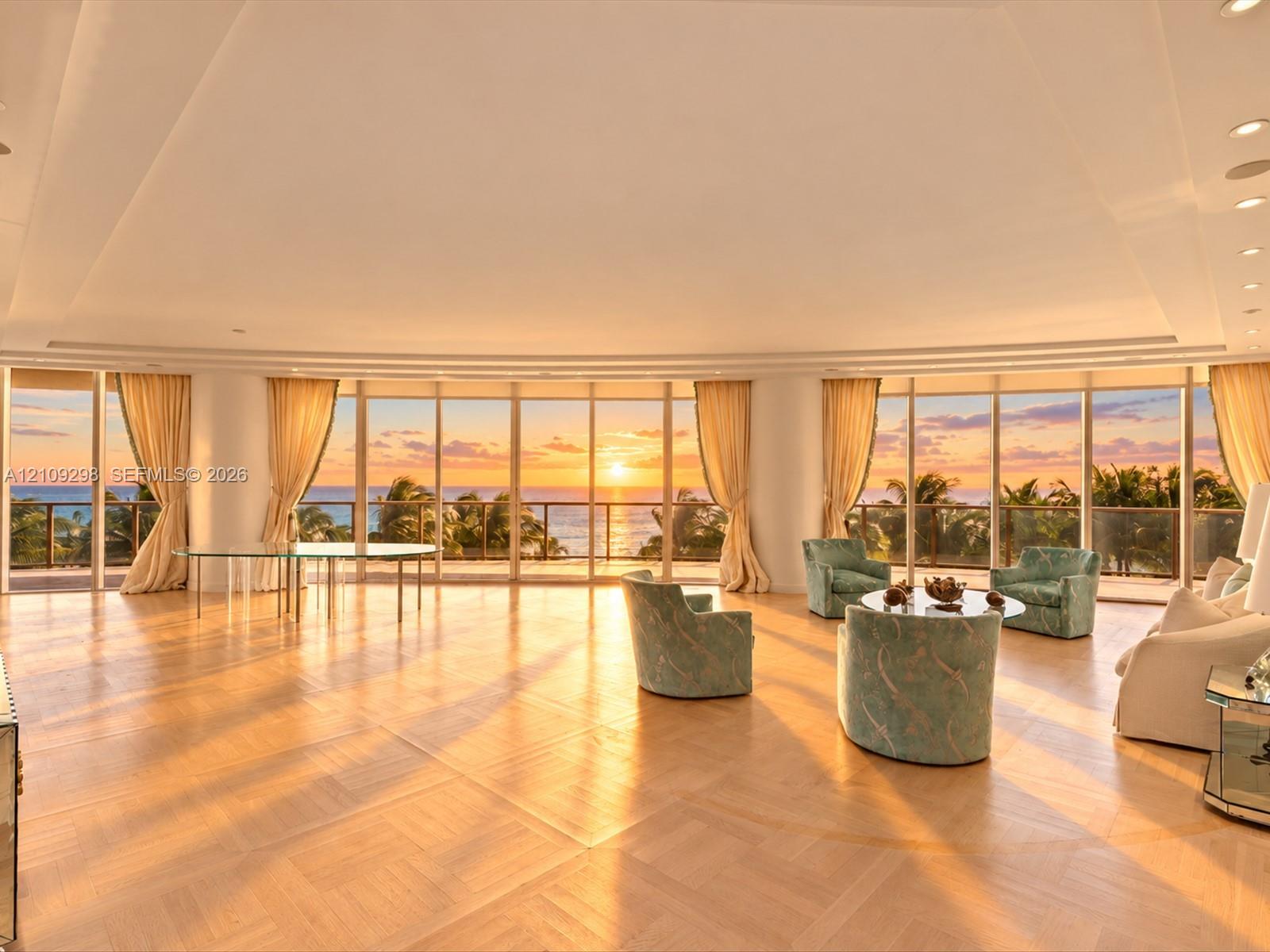 Curved floor-to-ceiling windows, herringbone-patterned light wood flooring, tray ceiling with recessed lighting, and a wrap-around balcony with ocean and horizon scenery