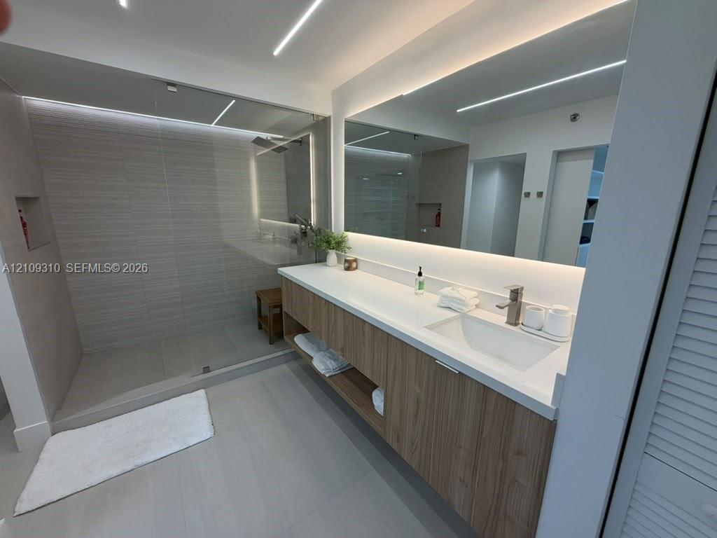 Floating wood-finish vanity with a white countertop and integrated sink, frameless glass walk-in shower with horizontal tile wall detailing, and a large-format LED-backlit mirror