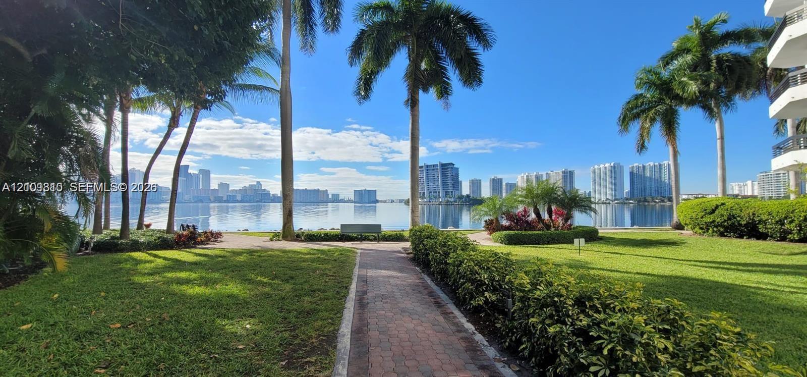 Waterfront grounds featuring mature palm trees, manicured lawns, paved walkways, and building structure with balconies