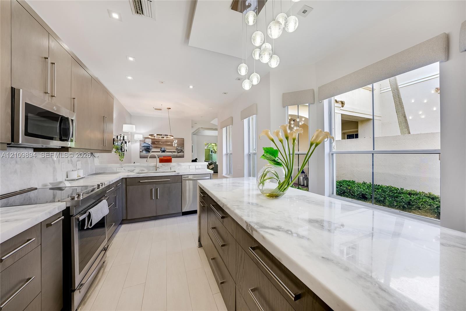 Light-toned wood-finish flooring, circular LED chandelier, textured wall covering, recessed ceiling lighting, and quartz-topped island with stone-finish waterfall edge
