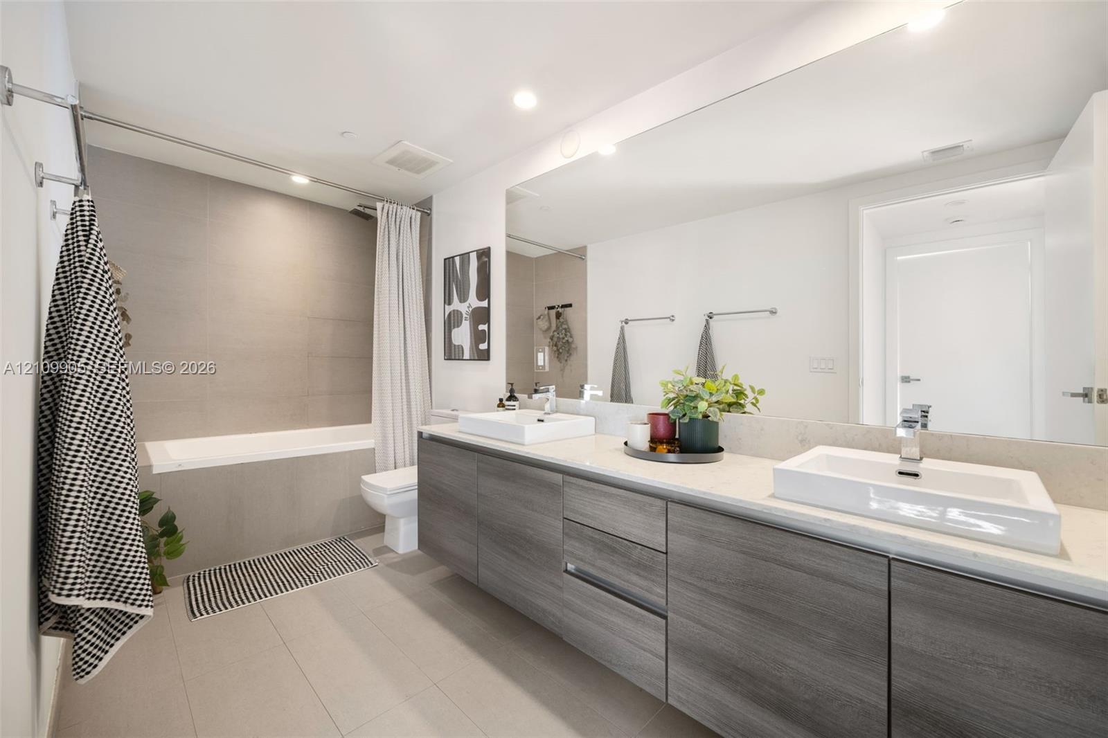 Double vanity featuring wood-finish cabinetry, quartz countertops, dual rectangular vessel sinks, and an expansive wall-mounted mirror, paired with a bathtub encased in neutral-toned large-format tile