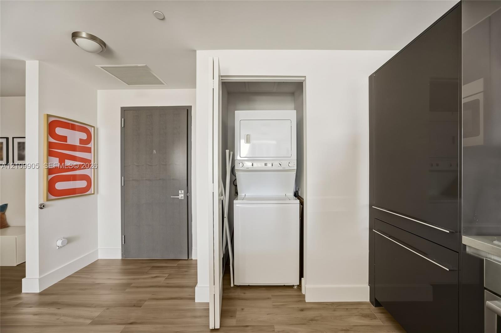 In-unit stacked laundry closet, solid core entry door with satin nickel hardware, high-gloss flat-panel cabinetry, and wood-finish flooring