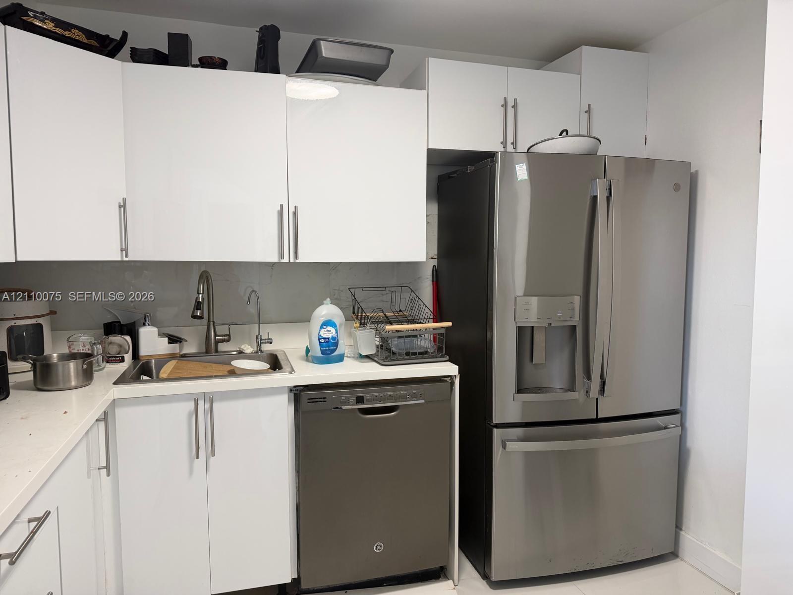 Modern kitchen area featuring flat-panel white cabinetry, white countertops, stainless steel French-door refrigerator, integrated stainless steel dishwasher, and a single-basin undermount sink with a high-arch pull-down faucet
