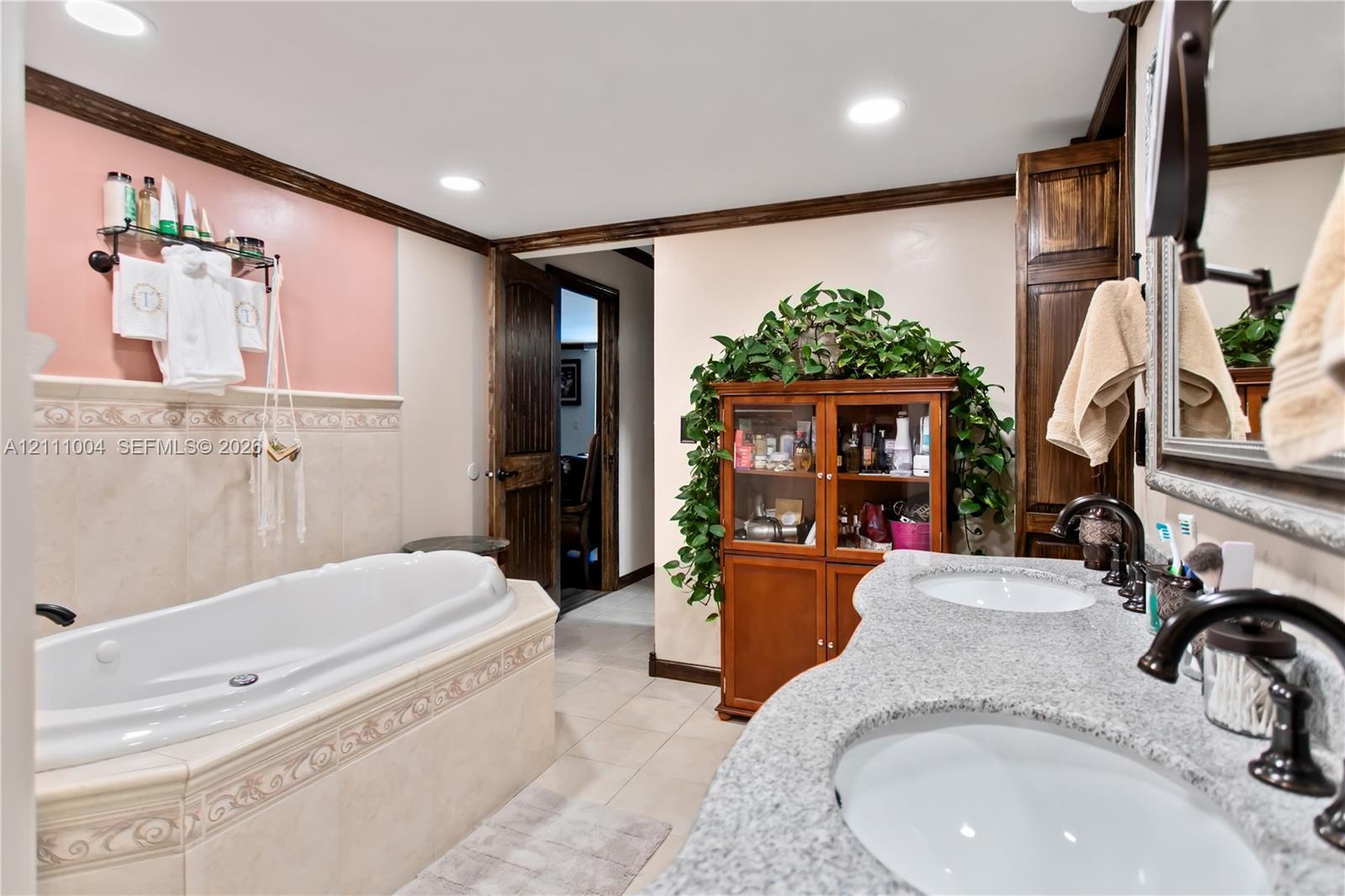 Dual-sink vanity with speckled stone countertop, jetted soaking tub with decorative tile surround, recessed lighting, and built-in wood cabinetry