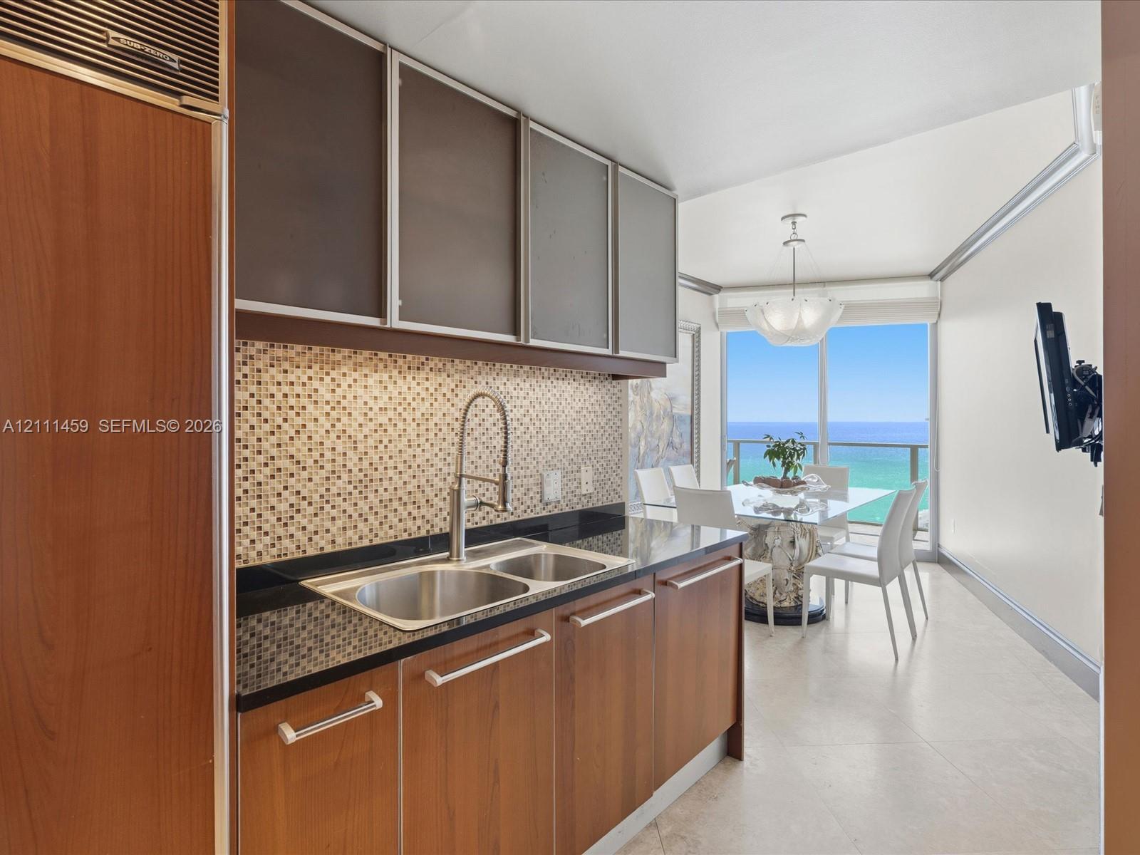 Kitchen featuring wood-finish cabinetry, a mosaic tile backsplash, dark solid-surface countertops, an integrated stainless steel refrigerator, and large-format floor tiling