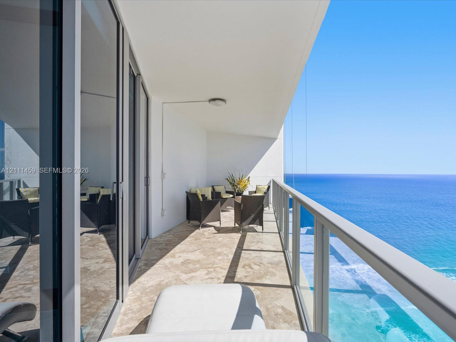 Covered balcony featuring stone-tile flooring, floor-to-ceiling glass doors, and a transparent glass railing overlooking the ocean