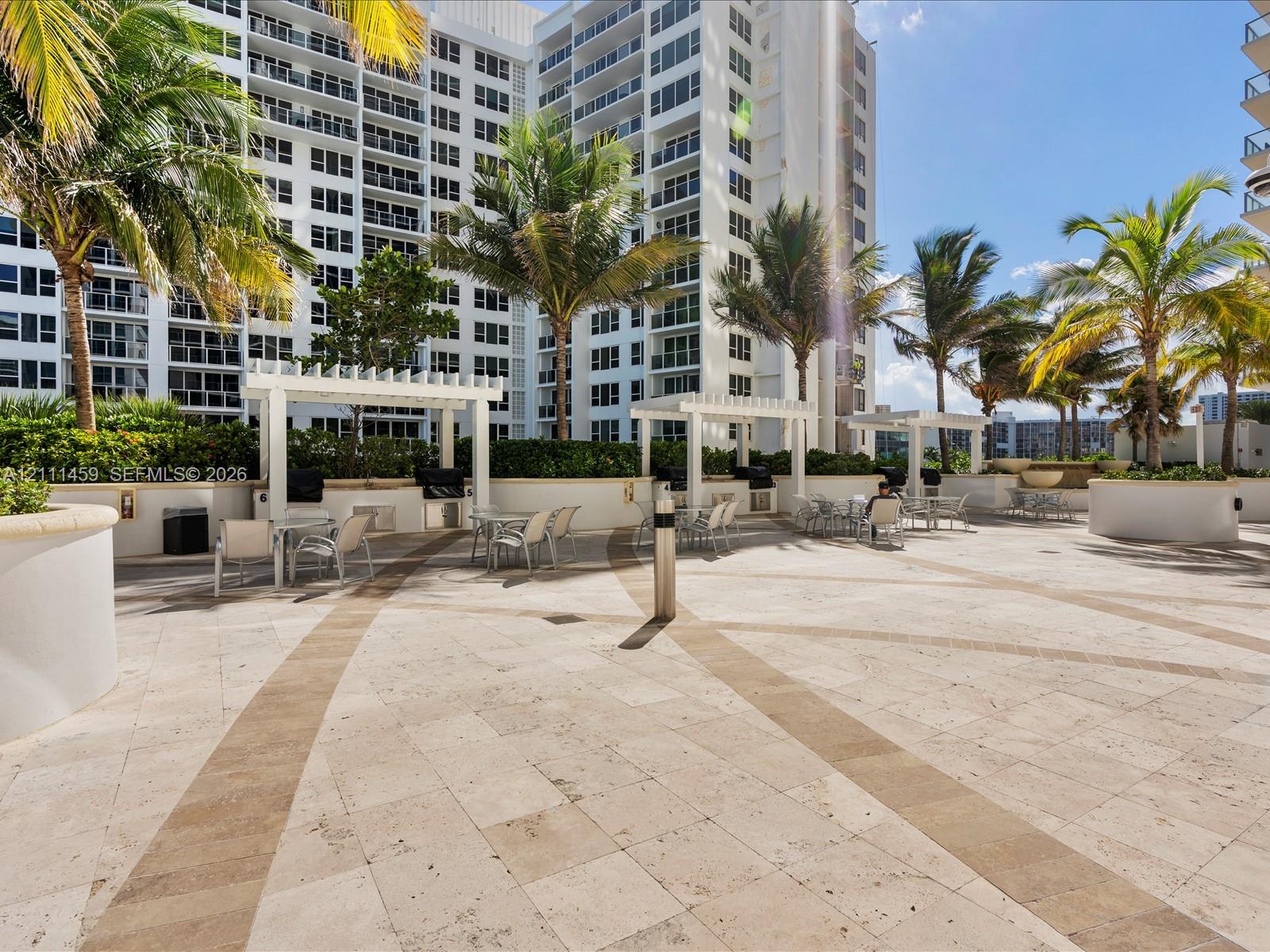 Stone-paved terrace featuring multiple white pergolas, built-in outdoor cooking stations, and mature palm landscaping