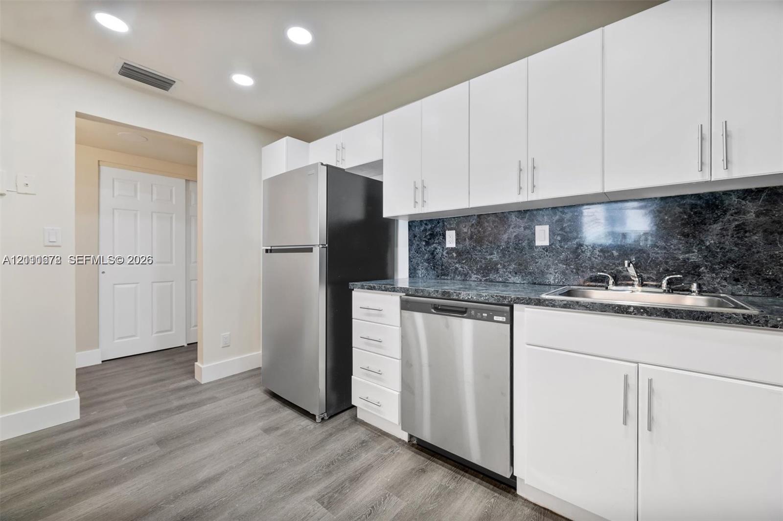 Kitchen featuring white shaker-style cabinetry, stainless steel appliances, dark stone-patterned countertops, a stainless steel sink with a high-arch faucet, and wood-finish flooring
