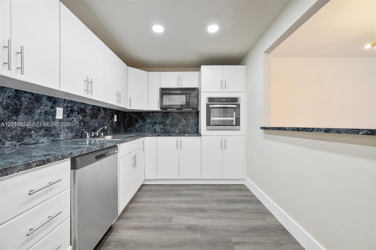 Kitchen featuring white shaker-style cabinetry, dark stone-patterned countertops, integrated stainless steel oven and microwave, stainless steel dishwasher, and wood-finish flooring