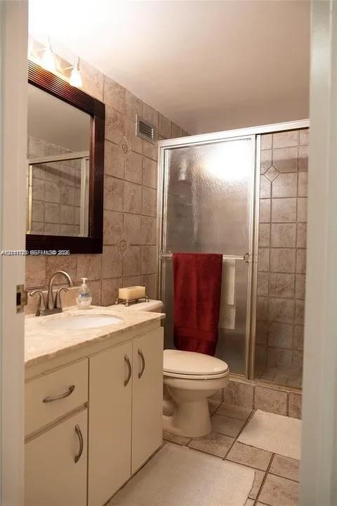 Bathroom featuring a vanity with a neutral countertop and integrated sink, a framed mirror, a walk-in shower with a glass door, and stone-finish wall and floor tile