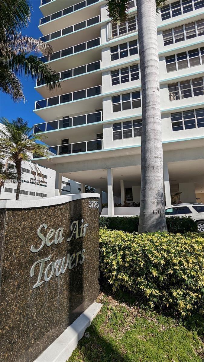 High-rise exterior featuring stacked balconies with glass railings, polished stone monument signage, and mature landscaping